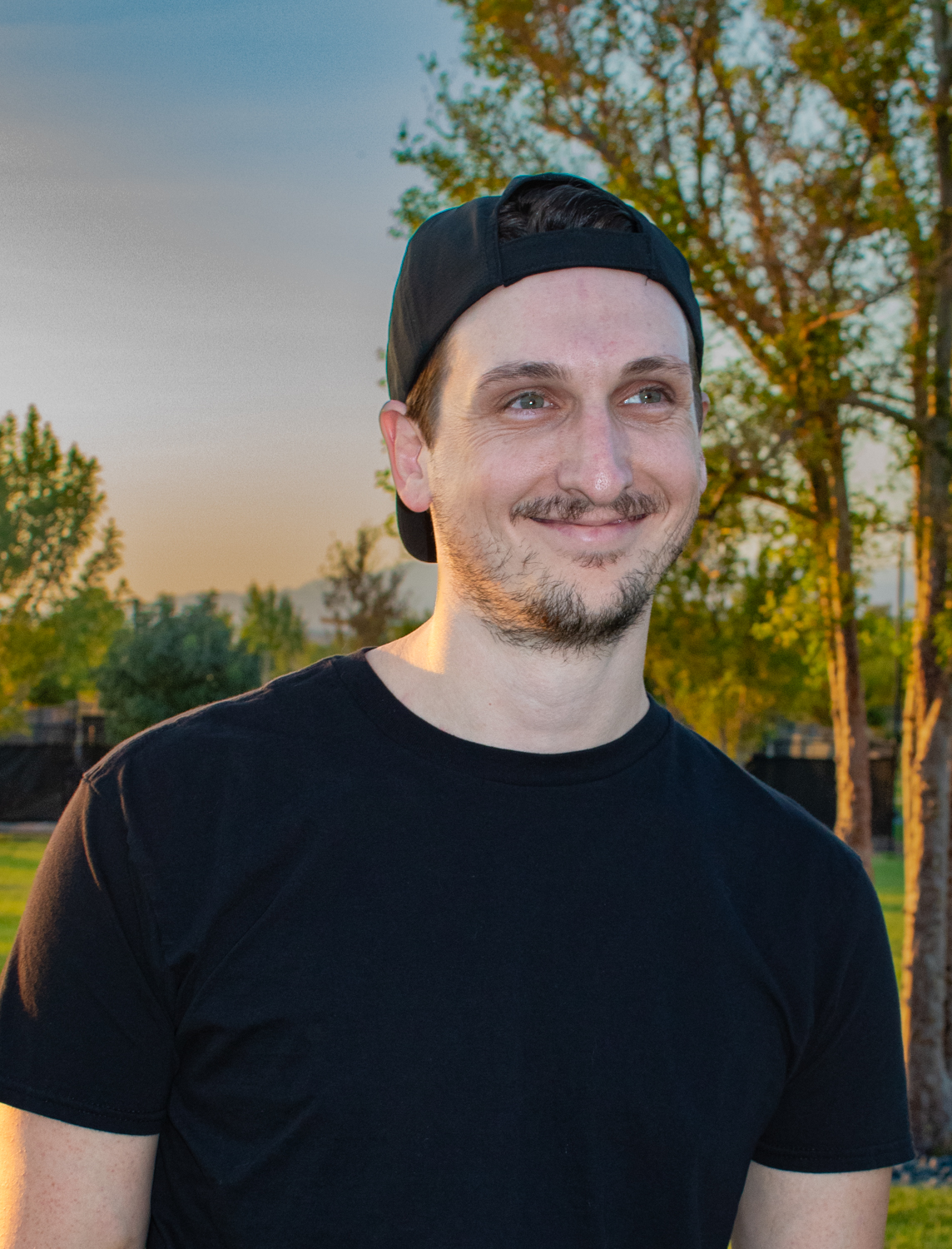 A man in a black shirt and a backwards baseball cap  smiles for a photo.