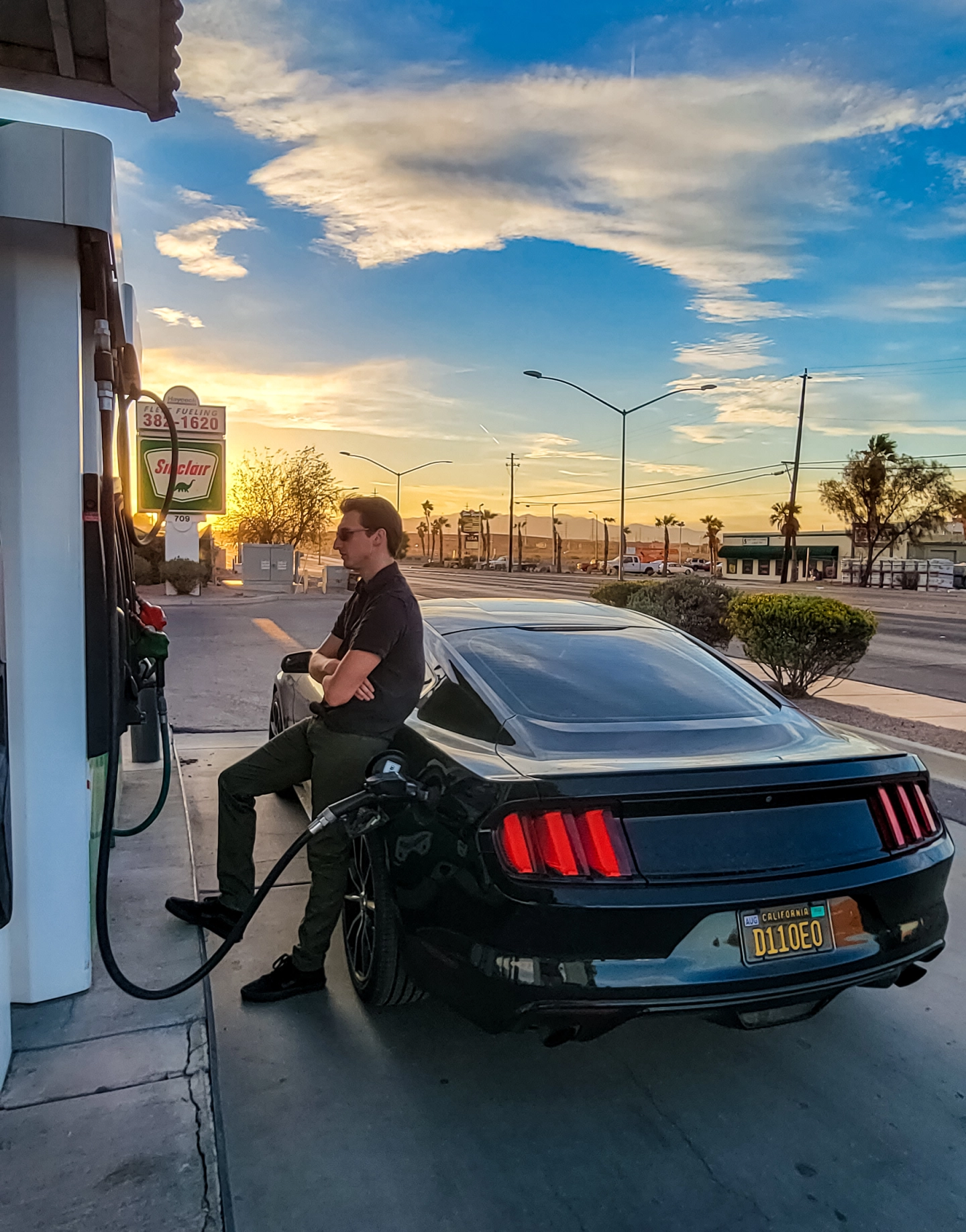 A man leaning against his car at the gas pump, with a sunset in the background